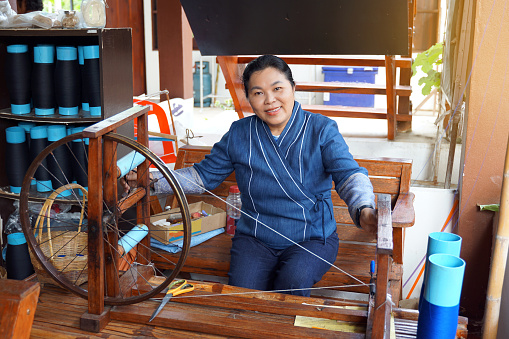 Thai women wearing traditional blue hand-woven clothes use a hand-winding machine. to prepare yarn for weaving in the concept of community products community enterprise Hand weaving, OTOP products