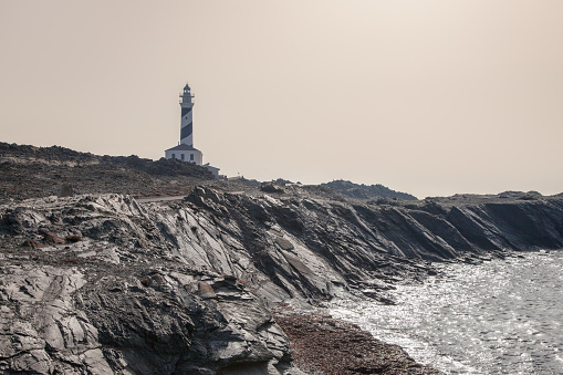 Explore the coastal allure in 'A Lighthouse in Menorca Island.' This captivating image captures the sentinel beauty of a lighthouse perched on a cliff near the sea. The maritime landscape of Menorca unfolds, merging architectural grace with the rugged charm of the cliffs. A mesmerizing portrayal of nautical tranquility.