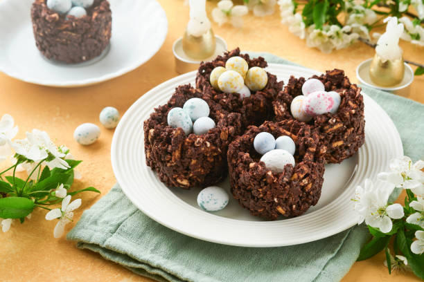 istock Easter chocolate nest cake with mini chocolate candy eggs with blossoming cherry or apple flowers on yellow background table. Creative recipe for Easter table with holiday decorations. Top view. 1795093265