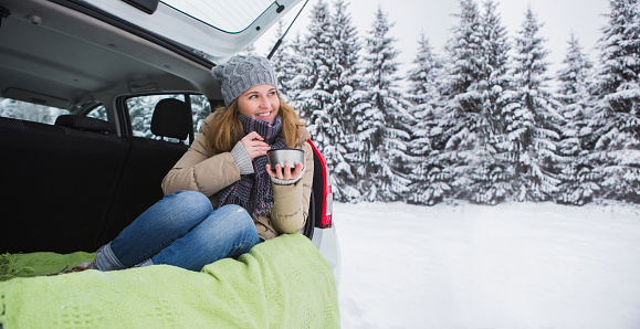 Woman in woolen hat sits in the trunk of the car and holds a cup of hot tea in her hands.