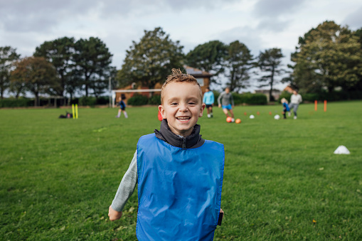 A medium, portrait shot of a young boy while playing football for a children's team in the North East of England. He is standing, looking and smiling at the camera on the sports field wearing a sports bib.