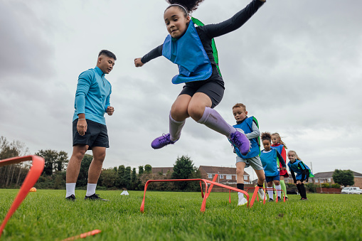 A low-angle, wide shot of a male football coach and a group of children wearing sports clothing, football boots and sports bibs on a football pitch in the North East of England. The coach stands and motivates his students as they run and jump over agility hurdles.