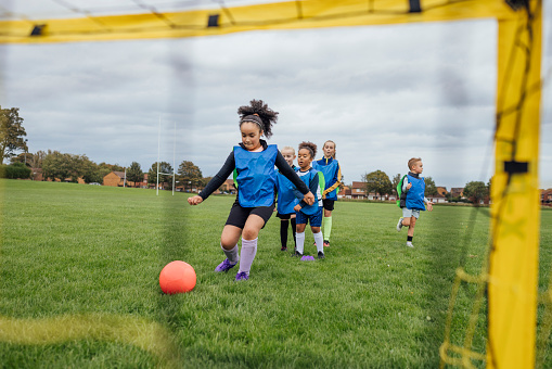 A wide shot of a group of children wearing sports clothing, football boots and a sports bib on a football pitch in the North East of England. They take turns shooting at the goal at football practice.