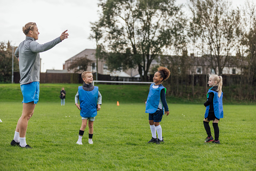 A wide shot of a male football coach and a group of children wearing sports clothing, football boots and sports bibs on a football pitch in the North East of England. He stands and coaches the children as they do football training drills.