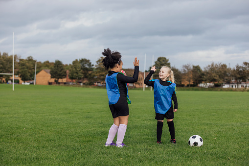 A wide shot of two girls wearing sports clothing, football boots and sports bibs on a football pitch in the North East of England. They stand in the middle of a football pitch together and give each other a high five.