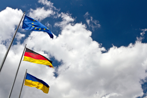 Three international flags of Ukranie, Germany and European Union against the blue and cloudy sky with a blank for copy space