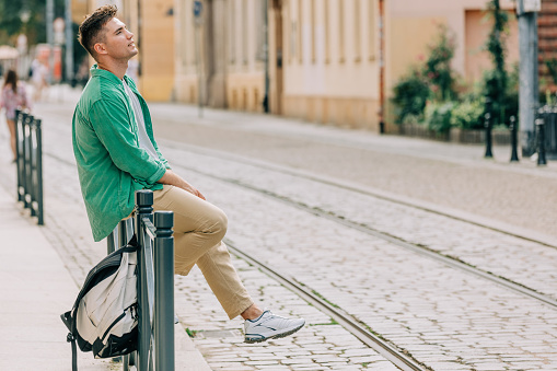 Stylish student guy in green shirt sits on railing on street