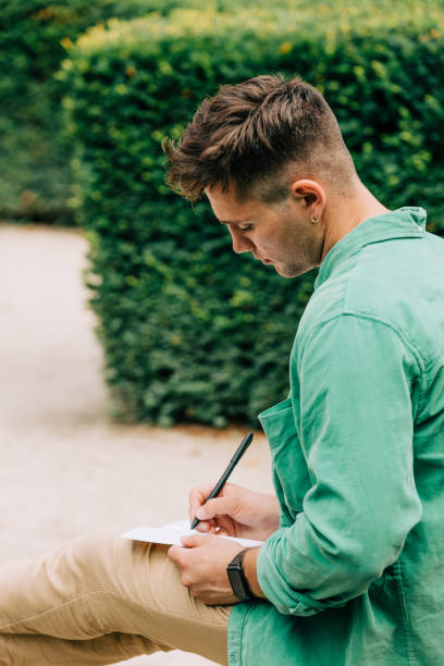 istock Stylish student guy in green shirt sits on bench in garden and write something 1795112238