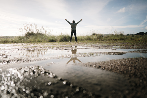 Rear view of a man with arms raised in a natural area, and his reflection on a puddle
