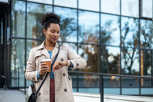 Portrait of Mature African American Entrepreneur Looks at Her Wristwatch in Front of Modern Business Building