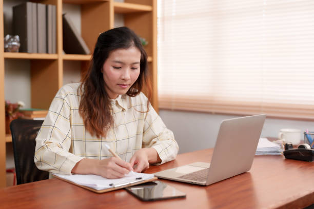 istock Office woman sitting working in front computer screen placed on table in front, eyes turned look at work information in document placed near computer, check work see if information matches or not. 1795160601