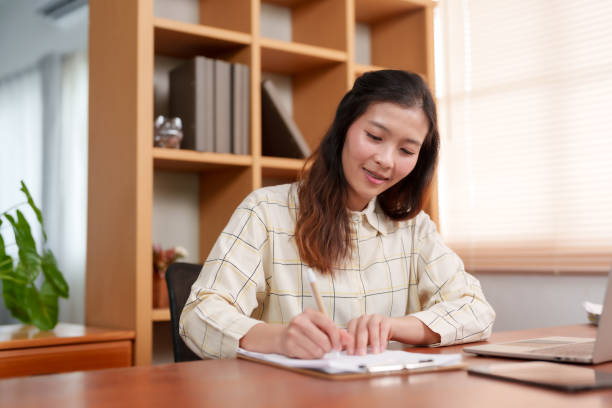istock Close-up, beautiful Asian office worker, sitting working on laptop computer placed on table in front, with carefree expression, reached out hand picked up pen write down main points work in notebook. 1795160612