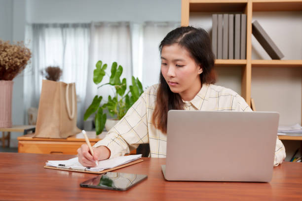 istock Asian woman, office worker writing work on sheet paper, for taking notes, but when message from friend came onto laptop, woman looked away from writing down paper answered message, with happy smile. 1795160632