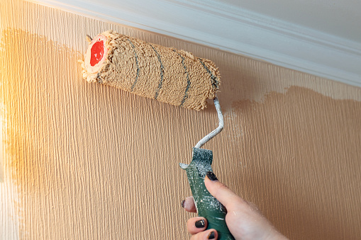 A woman's hand paints a wall with a paint roller in a beige color.