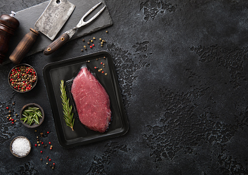 Raw beef fillet steak in vacuum tray with pepper on on black background with meat cleaver and fork.Top view.