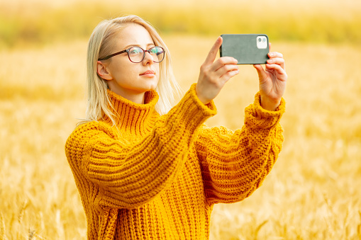 Stylish girl in eyeglasses and yellow sweater with mobile phone in wheat field