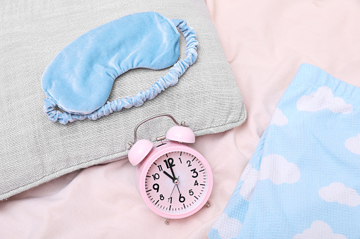 Light blue sleep mask, pillow and alarm clock on beige cloth, flat lay