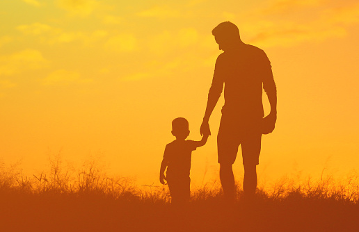 Father holding his son’s hand and walking in the meadow at sunset.