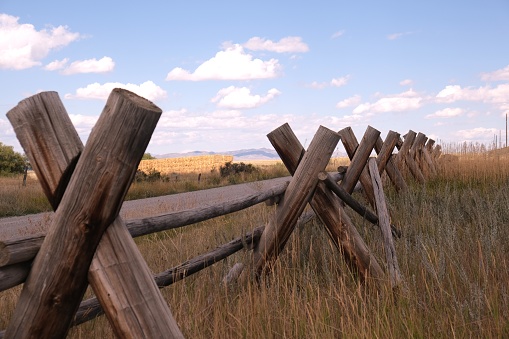 Various brown wooden split rail fences surrounded by green meadows and brown grasses in Wyoming with blue skies.  Deserted areas and property using various wooden rail fences for property lines and boarders with the sunlight shining.  Many photos with shadows from the rails in the sun and a couple with a pond showing the reflection of the cloud sky.