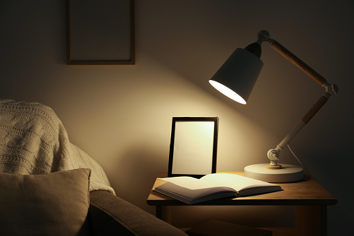 Stylish modern desk lamp, open book and frame on wooden cabinet in dark room