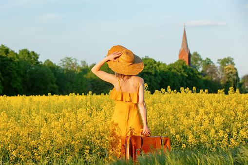 Stylish girl in yellow dress with suitcase in rapeseed field with village curch on background