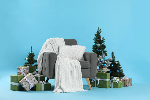 Beautiful Christmas themed photo zone with stylish armchair, trees and gift boxes on light blue background