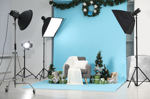 Beautiful Christmas themed photo zone with professional equipment, stylish armchair, trees and gift boxes in studio