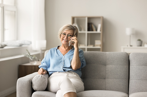 Positive pretty senior retired woman speaking on cellphone at home, enjoying conversation, leisure, mobile phone call on cozy couch, talking on cell, smiling, laughing