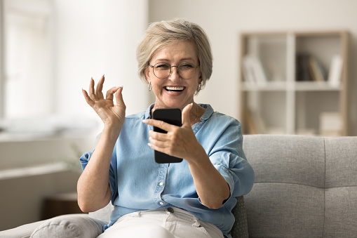 Positive cheerful pretty senior woman talking on video call on smartphone, showing OK hand gesture at mobile phone, enjoying communication, wireless connection, smiling, laughing