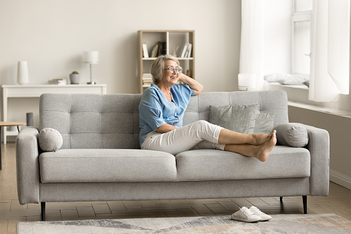 Cheerful dreamy senior pensioner woman relaxing on comfortable couch, outstretching barefooted legs, looking away, enjoying retirement, leisure at cozy modern home interior, smiling, laughing