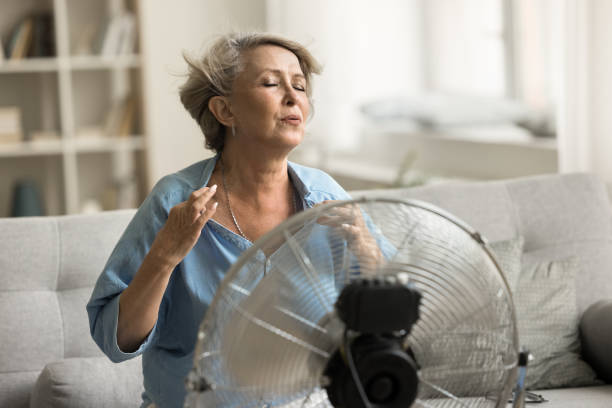exaustor mulher aposentada mais velha sentada em frente a um ventilador elétrico com hélice - menopausa fotos de stock