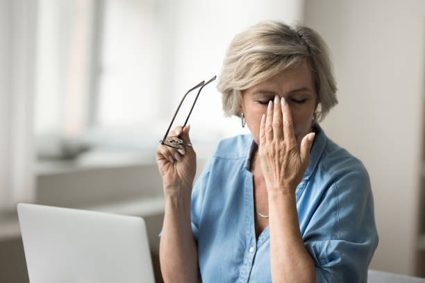 Tired elderly woman taking glasses off at workplace with computer