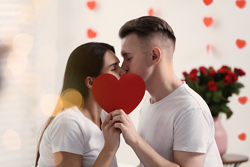 Lovely couple kissing behind red paper heart indoors. Valentine's day celebration