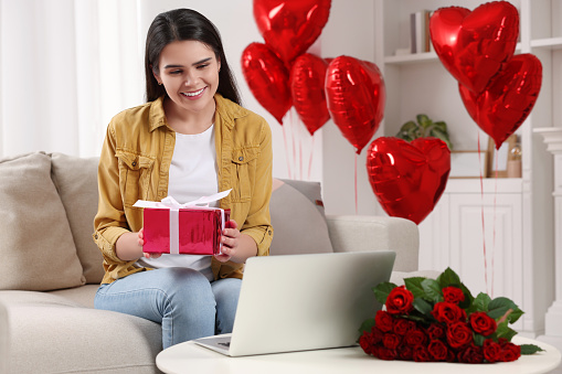 Valentine's day celebration in long distance relationship. Woman holding gift box while having video chat with her boyfriend via laptop at home
