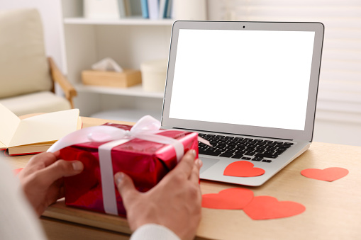 Valentine's day celebration in long distance relationship. Man holding gift box while having video chat with his girlfriend via laptop, closeup