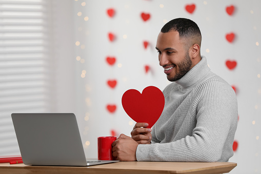 Valentine's day celebration in long distance relationship. Man holding red paper heart while having video chat with his girlfriend via laptop indoors