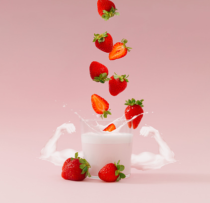 Creative layout made of strawberries falling into glass cup of milk drink with strawberry taste and arm muscles made of milk on pastel pink background. Minimal smoothie or milkshake concept.