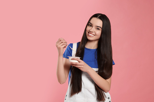 Happy teenage girl with delicious yogurt and spoon on pink background. Space for text