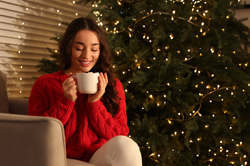 Beautiful woman with cup of tasty drink near Christmas tree indoors