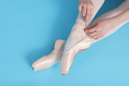 Young ballerina tying pointe shoes on light blue background, top view