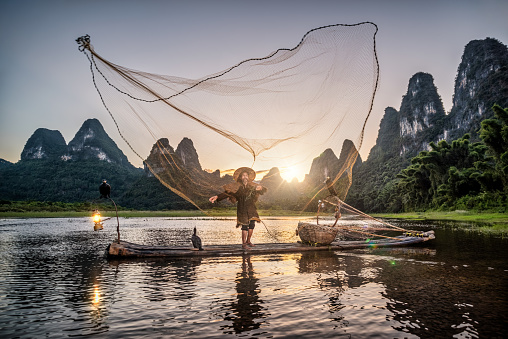 Traditional Chinese old senior fisherman in traditional clothes and bamboo hat on his wooden fishing raft with two cormorants fishing with a net on the Li River at sunset. Shot at Xing Ping, close to the city of Yangshuo County, Guangxi, Guilin, China.