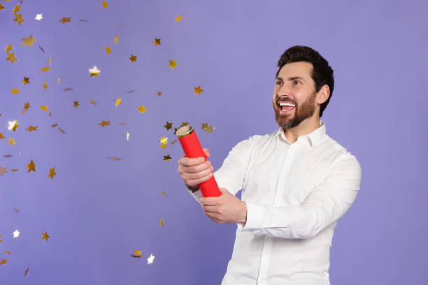 istock Happy man blowing up party popper on violet background 1795528004