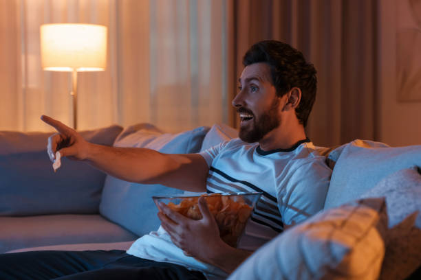 istock Man eating chips while watching TV on sofa at night. Bad habit 1795529059