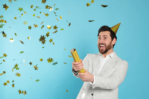Emotional man blowing up party popper on light blue background
