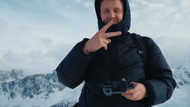 SLO MO Young Male Hiker in Hooded Jacket Operating Drone With Remote Control and Gesturing Peace in Snowy Dolomites