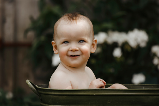 An adorable one~year~old baby boy smiles at the camera as he sits in an antique tub in a garden.