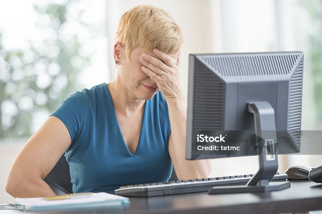 Tired Businesswoman Leaning On Computer Desk Tired mature businesswoman with hands on face leaning on computer desk in office Mature Women Stock Photo