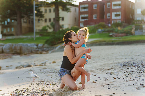 Beautiful multiracial woman of Pacific Islander descent kneeling in the sand at the beach and giving her cute toddler daughter a kiss on the cheek.