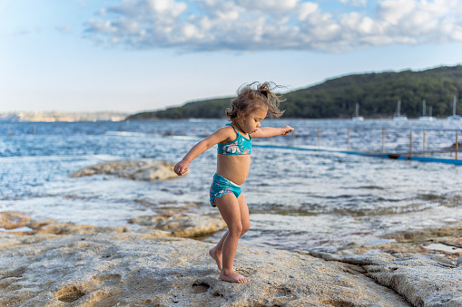 A cute Eurasian three year old girl wearing a swimsuit walks along the rocks while playing at a rock pool with her family while on vacation in Australia.
