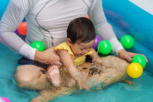 happy father and infant baby girl playing water splashing with colorful plastic balls in the inflatable pool